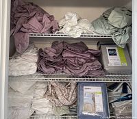 Shelf of folded bed linens including mauve, floral, sage green colors and packaged sheet sets from Sonoma and others.