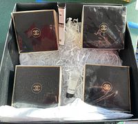 Top-down view of black gift box with shredded paper, containing four square black/gold Chanel boxes, one slender white sample box, and one small white tube.