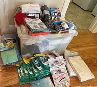 Photo showing a plastic bin filled with various cleaning cloths including Cuisinart branded towels, along with multiple packages of Dust Catcher cloths and Astonish Window Chamois cloths laid out in front of the bin.