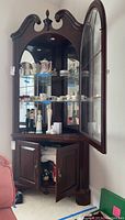 Front view of corner cabinet with open glass door showing glass shelves and displayed collectibles inside. Lower doors open showing storage inside.
