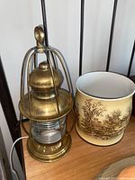 Photo showing brass lamp with cage-like structure and ceramic planter with brown scenic design