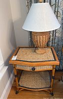 Rattan side table with drawer and lower shelf, topped with wicker base lamp and white shade.