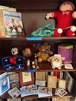Wide shelf view showing stuffed toys, Disney figurines in blue Swarovski cases, Furby toy in front of pink Furby box, brown teddy bear with blue ribbon, decorative glass item, and framed photographs on lower shelf.