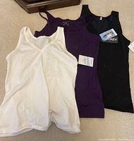 Three women's tank tops in white, purple, and black with the black one tagged as Jockey brand, displayed flat on carpet.