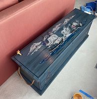Angled top-side view of wooden trunk showing painted sailing ship artwork, rope handles, and hinged lid.