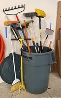 Large gray Rubbermaid BRUTE trash can filled with various yard and cleaning tools including brooms, mops, rake, and scrapers, with the trash can lid leaning against the wall