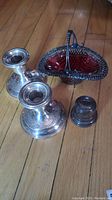 Two silver candlestick holders, silver-tone basket with red glass insert and ring box arranged on wood floor