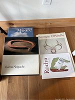 Four large art coffee table books arranged on wooden floor, featuring titles Celebrating Moore, Georgia O'Keeffe, Isamu Noguchi, and Rodin.