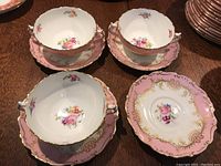 Three handled soup bowls each seated on matching pink and gilt saucers, additional saucers stacked right