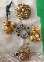 Photo showing seven brooches including a blue bird, gold-tone squirrel with faux pearl tail, pearl and rhinestone brooch, gold-tone rabbit with red eye, and rhinestone wreath brooch.