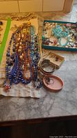 Photo showing multiple beaded necklaces in various colors laid on a surface, accompanied by a blue box with additional costume jewellery pieces inside including chains and earrings.