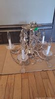 Photo showing the chandelier resting on the floor; five candle-style sockets with glass bobeches; multiple hanging glass crystal pendants in teardrop and bead shapes; silver-toned metal frame with curved arms.