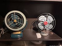 Two vintage fans side by side on a shelf, Torcan and Vornado.