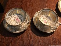 Two Paragon commemorative teacup and saucer sets displayed showing gold scrollwork, floral interior design, and coronation emblem