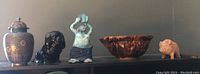 Full view of all five items on shelf: metal urn, black ceramic dog, blue-green figure, brown bowl, small tan dog