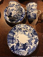 Photo showing 4 cups and saucers with 1 dessert plate on wooden surface, highlighting the Asian-inspired blue and white scenic pattern with gold trim.