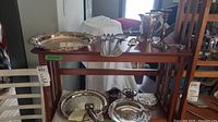 Overview of silver plated items on two wooden shelves including tray, teapot, candlestick, trivet, and spoons.