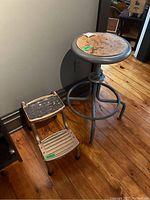 Photo showing metal stool with wooden round top and metal step stool with two steps on wooden floor near wall.