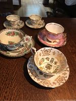 Five teacup and saucer sets displayed on a table, showing detailed patterns including gold coat of arms, floral, and pink gold trim sets.