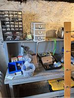 Wide shot of a workbench with assorted small containers and plastic drawer units holding screws, nails, bolts, and miscellaneous hardware parts, with toolboxes underneath and wood hooks on the right