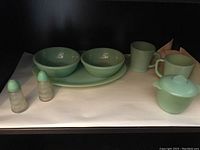 Set of 8 vintage Fire King Jadeite dishes displayed against a white and dark backdrop showing platter, two bowls, two mugs, covered sugar bowl, and salt and pepper shakers.