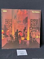 Front cover of ABBA The Visitors vinyl showing band members in dimly lit room with framed pictures on walls.