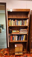 Front view of wooden four shelf bookcase with various books and a wooden box on bottom shelf (contents not included).