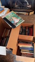 Cardboard boxes filled with assorted books including some hardcovers, paperbacks and children's books, showing overall worn condition.