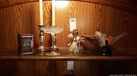 Lot on wooden shelf: two metal candle holders with beige taper candles, decorative tin box, paper angel figure, small woven basket, small stemmed glass vase with feathers, clear stemmed glass dish.