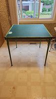 Front view of square green leather folding card table with metal legs, showing the flat playing surface and frame condition.