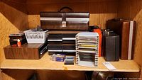 Shelf with multiple storage cases and bins including cassette and CD storage, portable leatherette case, and some cassette tapes.