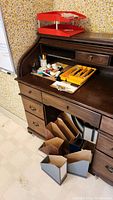 Photo showing wooden roll-top desk with multiple drawers, yellow desk drawer organizer with office supplies, red two-tier document tray on top of the desk, grey and brown magazine file holders below desk, cork board and whiteboard on wall.