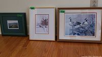 Three framed prints side by side on floor: smallest 5x4 landscape photo in blue-green frame, middle 6x8 deer print titled 'Regal Watch' by Dale Cooper in light wood frame, largest 12x9 Canadian geese print in similar frame.