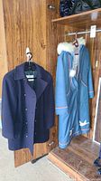 Both coats hanging: men's navy pea coat and women's blue trail coat with white fur trim and decorative embroidery