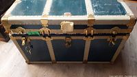 Front view of flat top trunk showing metal latch with keys attached, metal corner reinforcements, and blue panels.