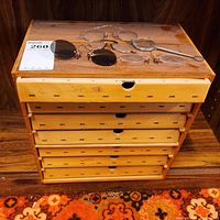 Top view of the wooden display box showing four vintage eyewear items placed on top: two pairs of metal round-lensed glasses, clip-on sunglasses lens, and magnifying glass. The box drawer fronts are labeled with years starting from 2000 to 2007.