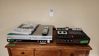 Two vintage electronics items on wooden cabinet: Realistic SCT-14 tape recorder and Panasonic VHS/DVD player with manual and remote on top