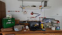Photo of a workbench area showing lawn care supplies, tools, a green crate with clothes pins, several small boxes, and household containers arranged on surface and shelves.