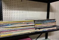 Two stacks of about 60 vinyl record albums on a metal shelf with pegboard wall background.