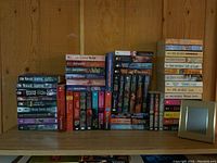 Collection of paperback novels stacked on a wooden surface against a wooden wall, showing many titles and colorful covers.