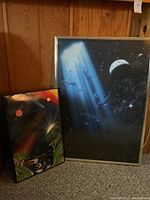 Two framed posters placed upright against wood paneling. Larger poster depicts underwater dolphins in cosmic space with planet and stars. Smaller poster shows landscape with planets and atmospheric light effects.