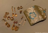 Photo showing all twelve pairs of varied styled gold toned earrings and the embroidered bag together on a surface.