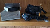 Photo showing vintage Sony Handycam and Sawyer slide projector with tray on a wooden surface along with a carrying bag and various small accessories including batteries and a small film reel.