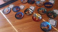 Twelve collectible plates arranged on a table, showing colorful illustrations from The Lion King movie including Simba, Nala, Timon, Pumbaa and other characters. Nine visible Certificates of Authenticity included with the plates. A black wall display rail is present next to the plates.