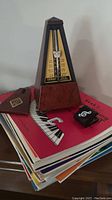 Wittner Maelzel mechanical metronome on top of a stack of assorted piano sheet music books. Metronome has a dark wood base and black plastic top with yellow scale and tempo markings.