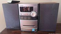 Front view of Sony Micro Hi-Fi Component System main unit flanked by two Sony speakers on each side, remote visible on top