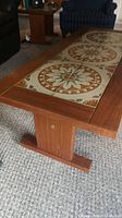 View of rectangular teak coffee table with four decorative patterned ceramic tiles inset in the top surface. The table is set on a carpeted floor with living room furniture nearby.