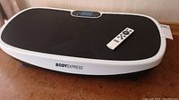 Black Body Express vibration exercise board with textured mat surface, digital control panel visible on one end; remote control placed on board.