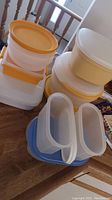 Stack of assorted Tupperware containers with orange and white lids on wooden table by railing.