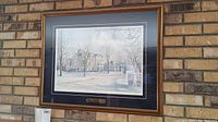 Full view of framed and matted print showing a winter scene of St Thomas courthouse with bare trees and snow, hanging on a brick wall.